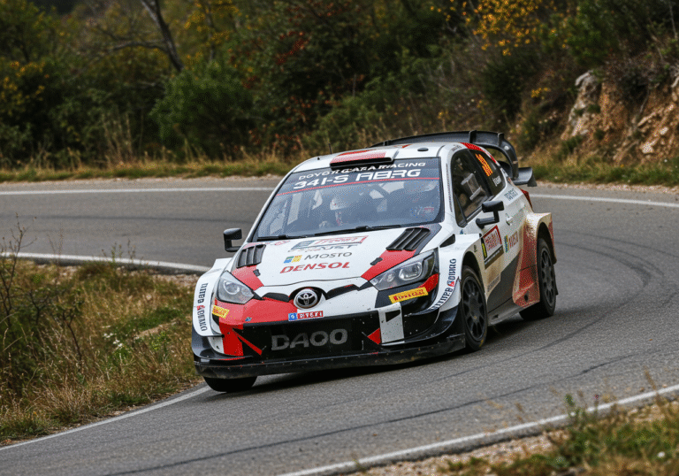 A focused action shot of Yuki Yamamoto, a TOYOTA GAZOO Racing WRC Challenge driver, intensely concentrated while navigating his rally car through a sharp turn on a winding asphalt road during the Croatia Rally. The car's tires grip the challenging Croatian road surface. Overcast natural light characteristic of an autumn day in early October. Sparse, dry grass and some green shrubbery line the roadside.