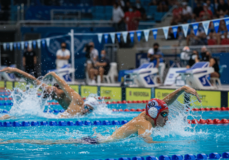 The Croatian men's 4x100 meter medley relay team, including Luka Čarapović, Filip Mujan, Vili Sivec, and Jere Hribar, in action during the World Aquatics Championships held in Singapore. One male swimmer, wearing a red and white Croatian swim cap, is powerfully pushing off the wall after a turn in the brightly lit indoor competition pool, with water splashing around him. Another teammate is visible in the water, swimming with intense focus. The background shows the professional setup of a large, modern indoor swimming arena in Singapore, with blurred spectator stands and strong, clear sports lighting. The atmosphere reflects the high-stakes global sporting event on its final day, August 3, 2025.