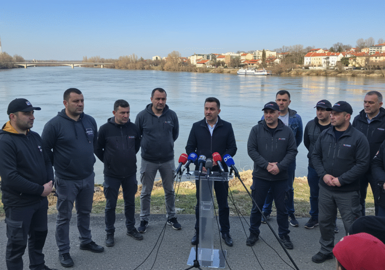A group of determined Croatian livestock farmers, led by Antun Golubović, addressing media outdoors at a press conference in Vukovar, Croatia. They stand near the confluence of the Vuka and Danube rivers, with the tranquil river waters and distant Vukovar buildings under a bright midday sun. The farmers, dressed in practical work clothes, show expressions of serious resolve and frustration, some with arms crossed or hands on their hips, conveying their strong demand for action from the Ministry of Agriculture. The scene is set against a clear blue sky, emphasizing the open and public nature of their protest announcement.