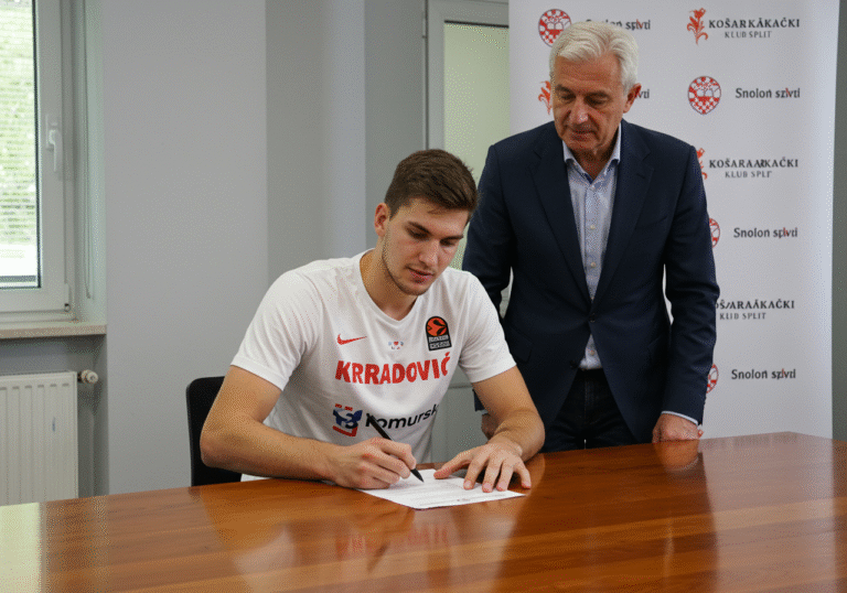 A 23-year-old Croatian male basketball player, Ivan Perasović, 200 centimeters tall, is seated at a polished wooden table. He is holding a pen and looking down at a document he is signing, with a focused and determined expression. A club official, an older man in a business suit, stands beside him, observing the signing with a professional demeanor. The setting is a well-lit indoor room at the Gripe sports complex in Split, Croatia. A subtle banner or backdrop with the Košarkaški klub Split logo is visible in the background. The atmosphere is formal and professional, characteristic of a significant sports transfer announcement. Natural indoor lighting.