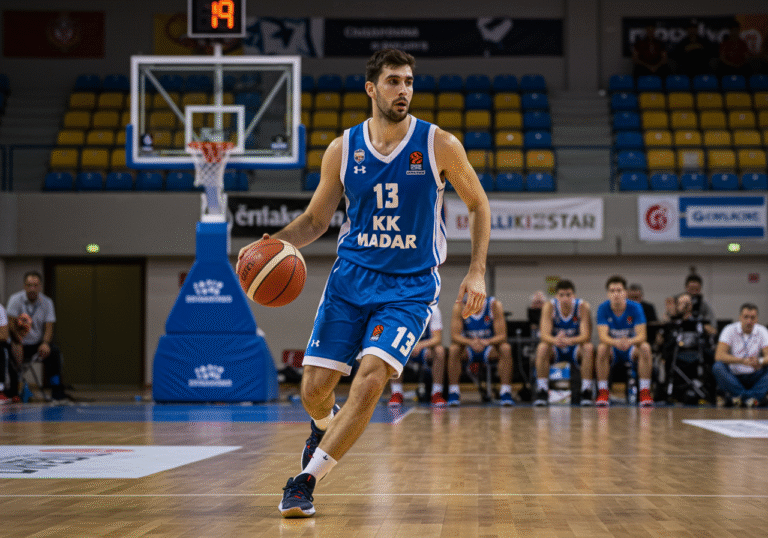 Ivan Vraneš, a 29-year-old male basketball player, dribbles a basketball on the polished hardwood court inside the Višnjik sports complex in Zadar, Croatia. He wears a blue and white KK Zadar basketball uniform. The bright, even indoor arena lighting illuminates the scene, showing a focused and determined expression on his face. Editorial sports photograph.