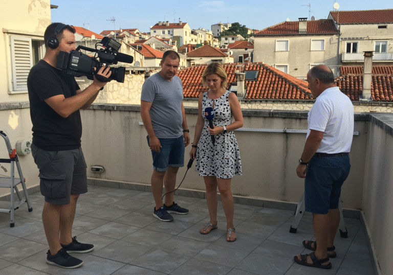 Danka Derifaj, a female journalist, Petar Janjić, a male cameraman holding a professional video camera, and a male citizen named Branko Miodrag are on a sun-drenched rooftop terrace in Žnjan, Split, Croatia, on a bright August day. One of them is in the act of stepping over a low partition fence or wall, with a small ladder used for access to the terrace visible nearby. The scene captures the residential architecture of Split and the clear summer sky in the background.