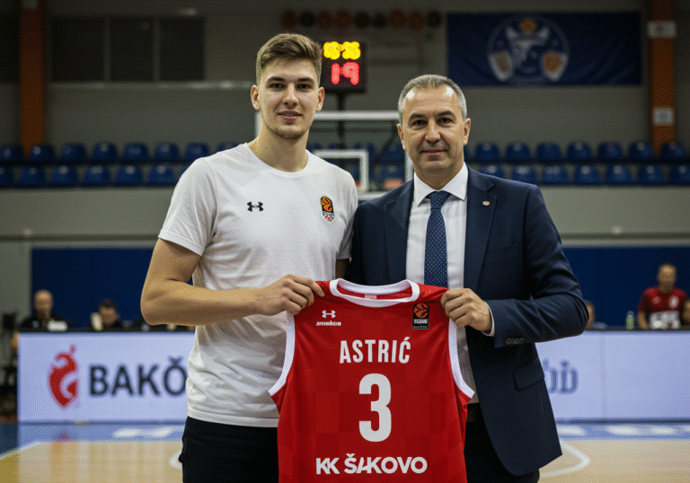 A 22-year-old Croatian basketball point guard, Ivan Ostric, stands in a well-lit indoor sports hall in Đakovo, Croatia. He is smiling slightly, holding up a new Košarkaški klub Đakovo (KK Đakovo) jersey with the club's emblem visible. A professional club official, possibly a coach or director, stands beside him, shaking his hand, both looking towards the camera. The background shows blurred elements of a basketball court or club facilities, typical for a formal player presentation.