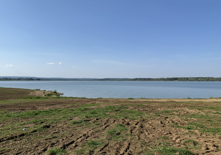 A wide shot of Jezero Čiče, a lake near Zagreb, Croatia, on a sunny day. The foreground shows an expanse of recently mowed and cleared ground along the shoreline, with some residual natural vegetation. The calm waters of the lake stretch into the midground, reflecting the clear blue sky. In the distance, subtle hints of the surrounding Croatian landscape, conveying a natural area undergoing preparatory work for a future recreational and bathing zone.