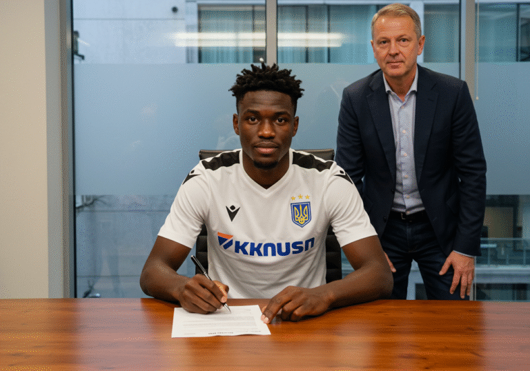 A professional footballer, Peter Itodo Mandela, seated at a desk in a modern, well-lit office, holds a pen and intently signs an official contract. Across from him, a representative of the Ukrainian football club Metalist 1925 is present, observing the signing. The setting is formal and concise, emphasizing the completion of a significant transfer deal, typical of a press conference or a private signing ceremony.