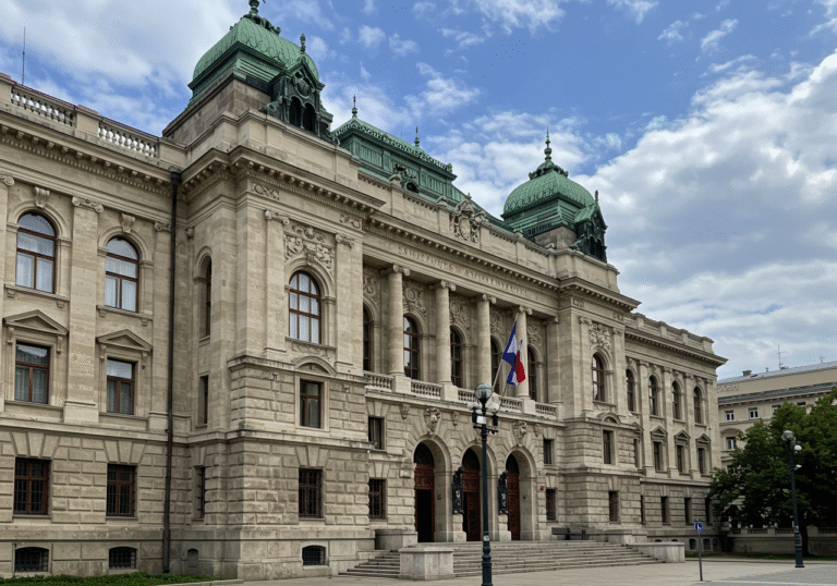 The stone facade of the Supreme Court of the Republic of Croatia building in Zagreb under a partially cloudy late afternoon sky. The grand structure stands quiet and still, its architectural details prominent, conveying the institutional gravity of the country's highest judicial body. The atmosphere is calm, reflecting the ongoing institutional blockade.