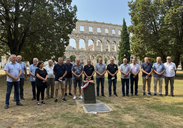 A daytime scene in Valerijin park, Pula, Croatia, on a bright summer Thursday. A respectful gathering of Croatian citizens, including members of the iconic rock band Parni Valjak and representatives from local Pula organizations, stand together in the park. Their expressions convey quiet remembrance and shared purpose. In the background, the grand, ancient Roman Pula Amphitheatre (Arena) rises under a clear blue sky, partially framed by the park's trees. Natural light illuminates the scene, capturing the community's support for the monument initiative.