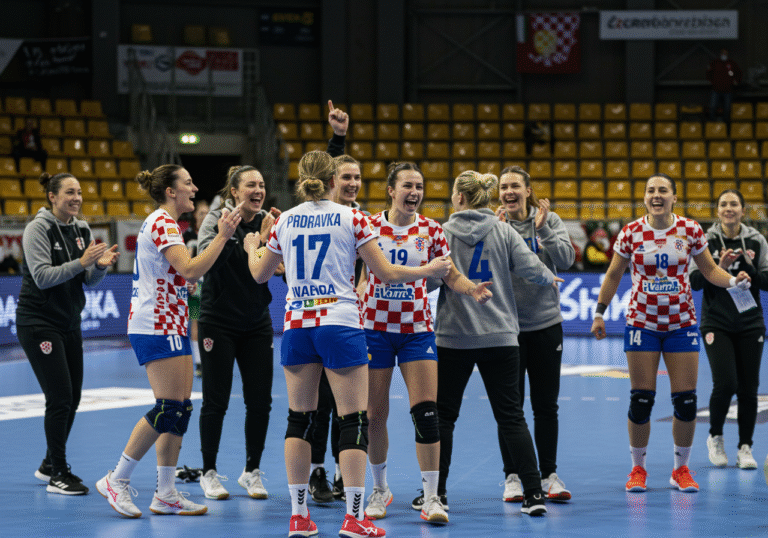 Croatian women's handball team Podravka celebrating their championship victory on the court after winning the Caring International Cup. Players, including Kristina Prkačin and Ana Panžda, are embracing and raising their arms in triumph in their team uniforms. The scene is set inside a modern sports arena in Székesfehérvár, Hungary, under bright indoor lighting, with a handball court visible. The atmosphere is one of exhilaration and collective achievement.