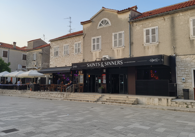 Daylight, wide shot of the exterior of Saints & Sinners nightclub in Poreč, Croatia. The entrance area appears quiet under a clear summer sky, with a subtle presence of Croatian police officers or faint yellow crime scene tape visible, indicating an ongoing investigation. The building displays typical Istrian coastal architecture. The overall atmosphere conveys the serious aftermath of a recent incident, rather than a lively club environment.