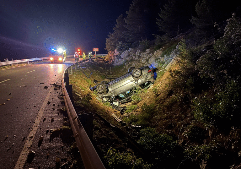 A nighttime view of a severe car accident on Krk island, Croatia, near Dobrinj. An overturned and heavily damaged passenger vehicle lies at the base of a steep, vegetated seven-meter ravine. On the winding road above, a section of the metal guardrail is visibly broken and bent, and surrounding vegetation is disturbed where the vehicle left the roadway after a curve. Flashing emergency lights, casting red and blue hues, illuminate the wreckage and the challenging, rugged terrain from the roadside, creating a somber atmosphere.