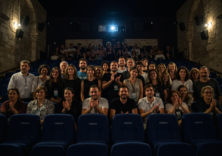 A diverse group of film festival attendees and organizers gathered inside a darkened screening hall at the Croatian International Film Festival (CIFF) in Šibenik, Croatia. Their faces are brightly illuminated by the glow of a large screen, showing expressions of enthusiastic delight and warm reception. Some individuals are smiling broadly, others are intently watching the projected image, with a few beginning to applaud. The ambient light is low, characteristic of a cinema, creating a focused atmosphere. The setting subtly reflects the historical and cultural architecture of Šibenik, blended with modern festival elements, capturing the excitement of a significant cultural event.