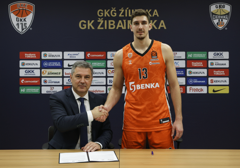 A realistic depiction of Petar Vujacic, a 25-year-old, 196cm tall Slovenian basketball player, in a formal signing moment. He is dressed in a GKK Šibenka basketball jersey, featuring the club's distinctive orange and black colors, and is shaking hands with a club official in a suit. A signed contract rests on a table in front of them. The background shows an interior setting, likely a press room or a club office within a sports facility in Šibenik, Croatia, with subtle club branding visible. The lighting is bright and even, characteristic of an official photo opportunity, capturing a moment of anticipation and professionalism.