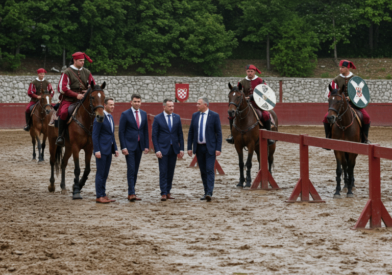 A wide shot capturing the rain-soaked, muddy track of the Sinjska alka tournament in Sinj, Cetinska krajina, Croatia. Several Sinjska Alka knights in their traditional historical uniforms are mounted on their horses, looking serious or concerned as heavy rain visibly wets their attire. Officials from the Viteško alkarsko društvo, including President Stipe Jukić, stand near the track, their expressions somber as they assess the dangerous conditions. The sky is overcast, and the ground is visibly slick and waterlogged, emphasizing the reason for the postponement of the event.