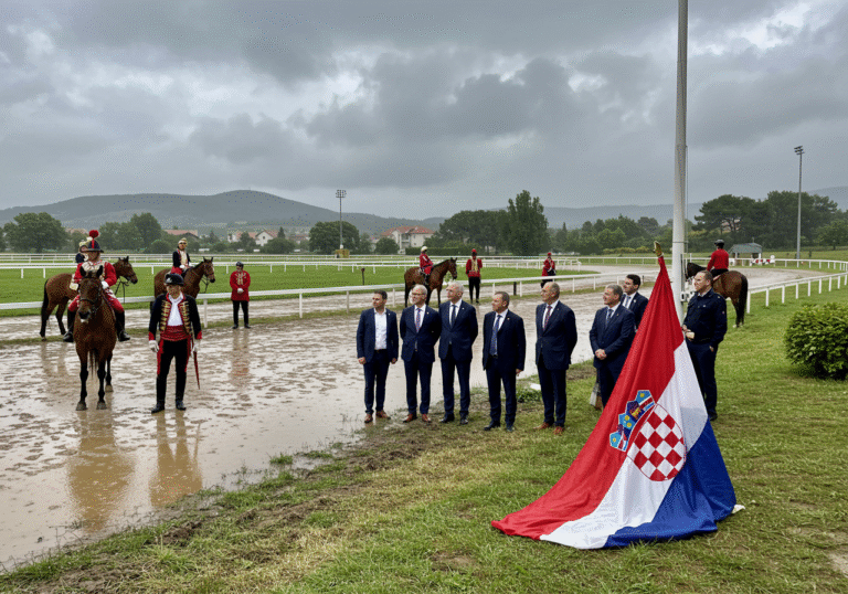 An outdoor scene capturing the Alkarsko race track in Sinj, Croatia, during a severe mid-afternoon thunderstorm. Torrential rain pours down, creating widespread puddles and turning the dirt track into deep, treacherous mud. Overcast skies are dominated by dark, ominous clouds, with faint flashes of lightning visible in the distance. Several Alkari, in their traditional elaborate uniforms and plumed helmets, stand by the saturated track, their expressions somber with disappointment. Some Alkari horses are being carefully led away towards nearby shelters. High-ranking Croatian state officials, some holding umbrellas, are observing the challenging conditions, their faces serious. A large, rain-soaked Croatian flag hangs heavy and limp from a pole near the track, reflecting the dramatic mood of the postponed event.