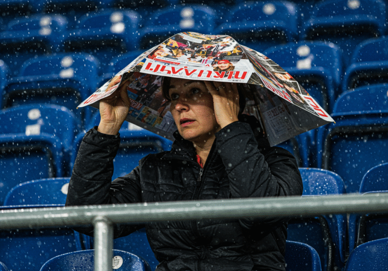 Sonja Jandroković, captured candidly in the spectator stands at the Sinjska Alka in Sinj, Croatia, during a sudden, heavy daytime downpour. She is using an ordinary magazine as an improvised umbrella, shielding herself from the rain visibly falling around her and hitting the stands. Overcast light, a sense of immediate adaptation to the extreme weather.