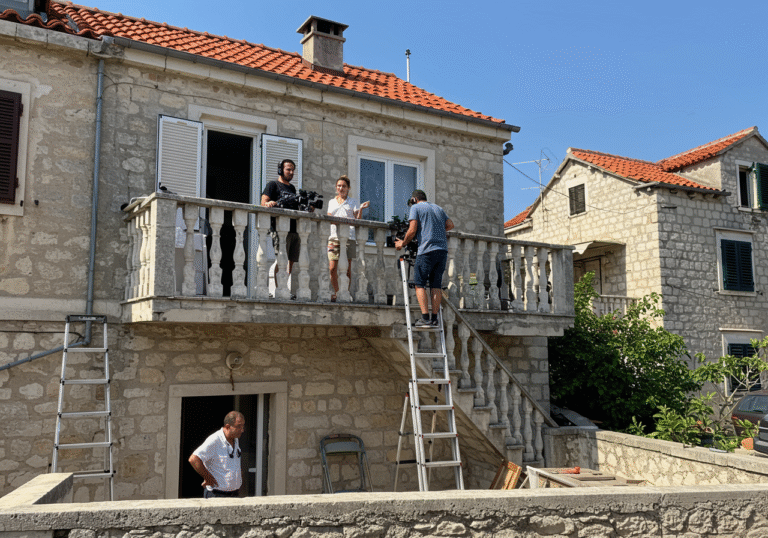 A bright, sunny August daytime scene in Split, Croatia. On the elevated terrace of a typical Mediterranean-style family house, journalist Danka Derifaj Mikulaš is visible, along with cameraman Petar Janjić, who is holding a professional video camera. A sturdy ladder is propped against the exterior of the house or against a fence leading up to the terrace. In the immediate vicinity, neighbor Branko Miodrag stands, observing the situation. The architecture is characteristic of coastal Croatia, with clear blue skies overhead.